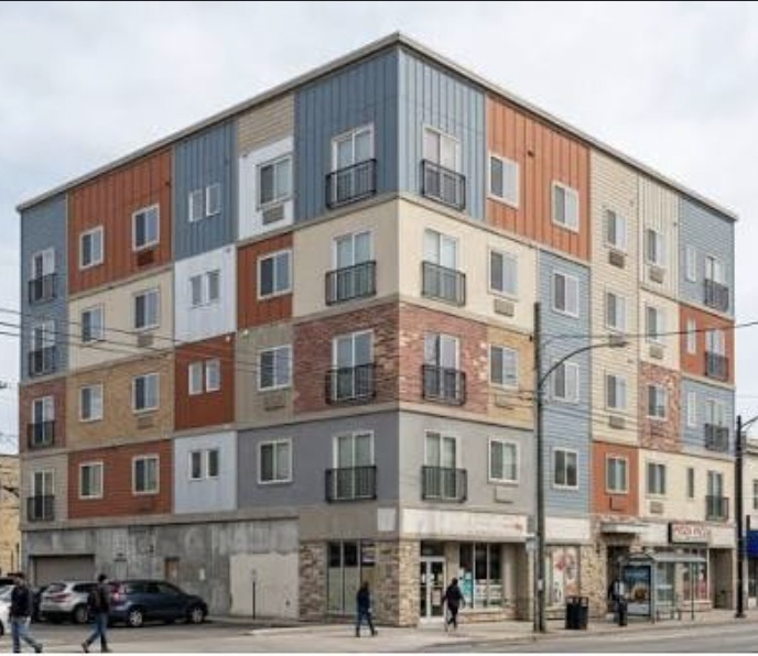 A typical five-story 5-over-1 apartment building on a street corner, with ground-floor retail and a patchwork of gray, blue, orange, and beige siding panels.